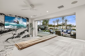 Quarto com uma cama grande, ventilador de teto, TV exibindo um surfista e vista para um pátio exterior com móveis.
