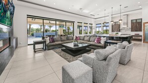 A spacious living area with a large sectional sofa, a coffee table, and a fireplace. There is a kitchen area with modern appliances and a dining table. - Palm Villa- Luxury 7 Bed, Pickleball, Volleyball, Golf (Indio)