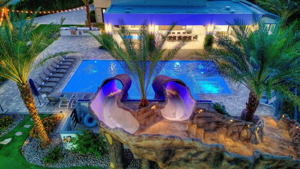 A pool area with two large rock formations, a lounge area with chairs and tables, and a building with a blue-lit exterior.