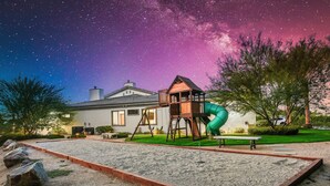 A playground with a slide and swings, a house in the background, and a starry night sky.