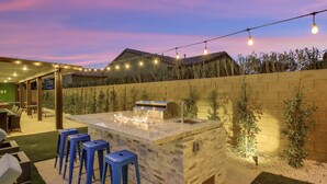 Uma área de bar ao ar livre com bancada de pedra, banquetas azuis e luzes de cordão.