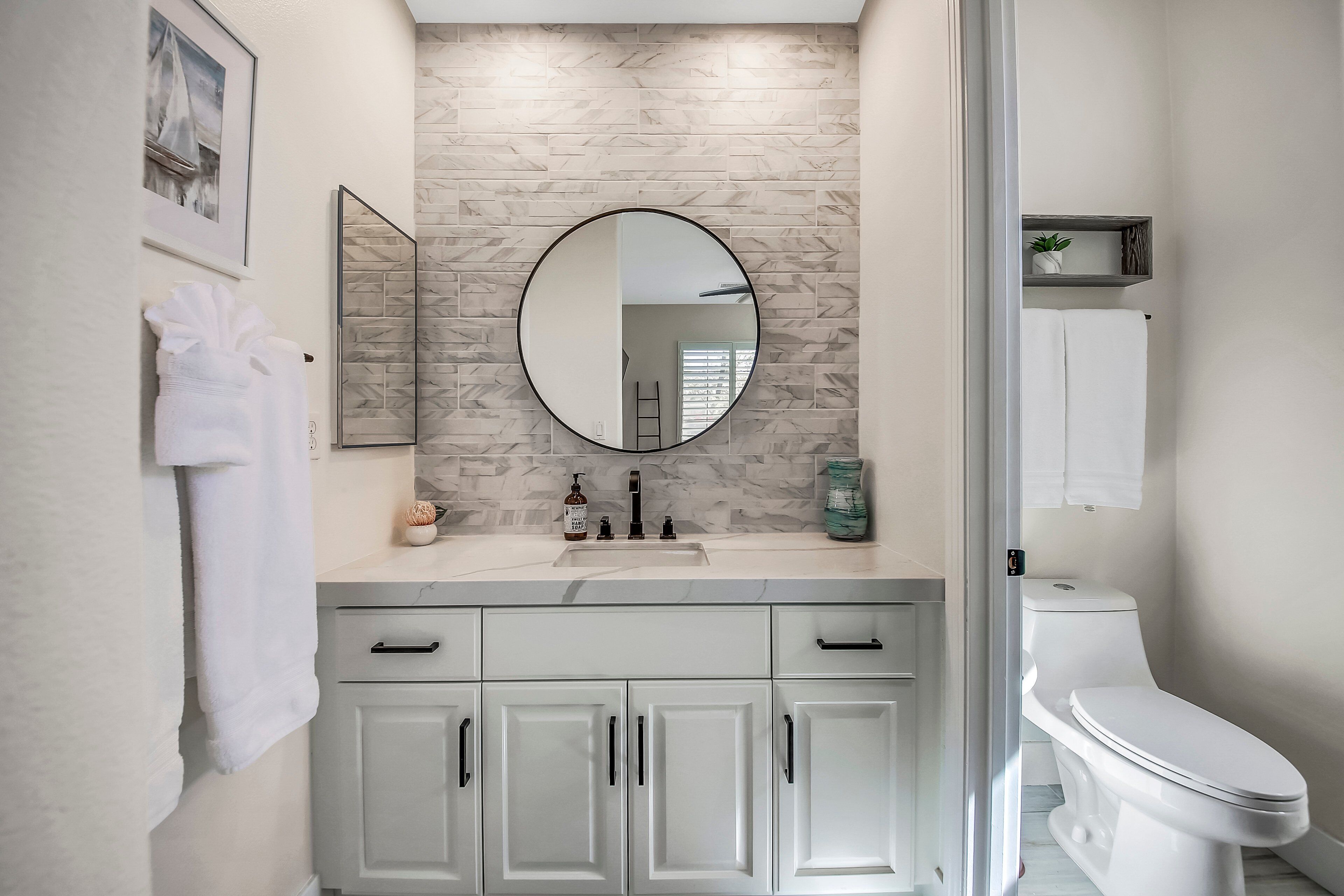 A modern bathroom with a marble backsplash, a round mirror, a white toilet, and a vanity with cabinets.