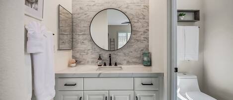 A modern bathroom with a marble backsplash, a round mirror, a white toilet, and a vanity with cabinets.