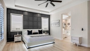 Quarto moderno com uma cama grande, mesas de cabeceira, ventilador de teto e vista para o banheiro.