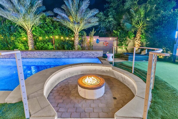A swimming pool with a fire pit in the center, surrounded by a brick patio and palm trees.