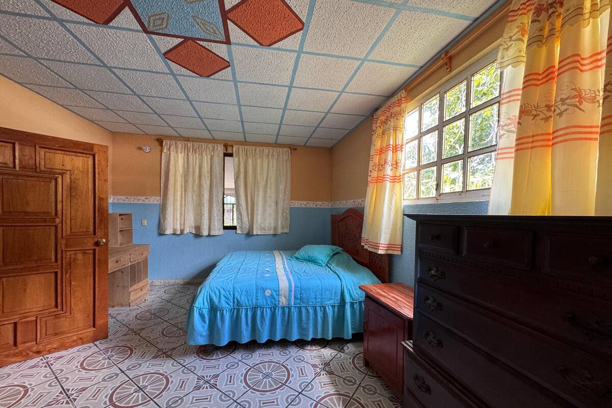 A bedroom with a patterned floor, a bed with a blue bedspread, a wooden dresser, and a window with curtains.