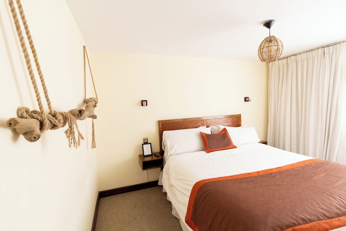 A hotel room with a large bed, a wooden headboard, a bedside table with a lamp, and a wall-mounted rope decoration.