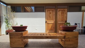A wooden bench with two terracotta planters on either side, set on a patterned tile floor in a courtyard with a white wall and wooden doors in the background. - Hostal Geosol (Antofagasta)