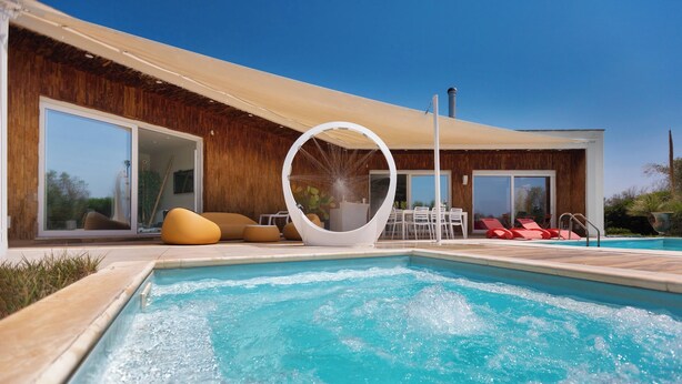 Un área de piscina exterior moderna con un elemento acuático circular, sillas de descanso y una terraza de madera.