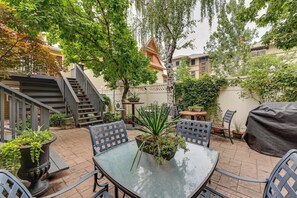 Un patio con una mesa de vidrio rodeada de sillas, plantas en macetas y una parrilla para barbacoa cubierta.