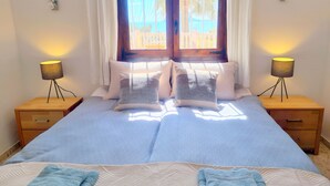Une chambre à coucher avec un lit, deux tables de chevet, une fenêtre avec des rideaux et une vue sur des palmiers.
