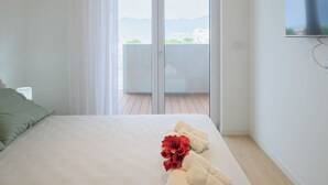 A hotel room with a bed, a TV mounted on the wall, and a balcony with a view of the city and mountains. - Porta del Mare Luxury Apartment (Salerno)