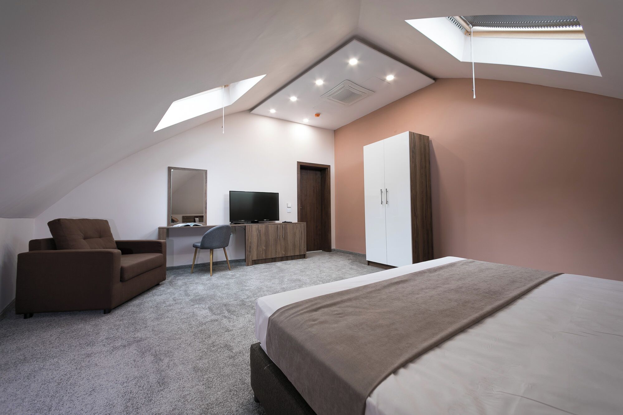 A modern bedroom with a sloped ceiling, a large bed, a desk with a chair, a wardrobe, and a television.