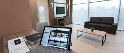 A laptop on a desk with a brochure, a sofa, a coffee table, and a TV in a modern living room with large windows.