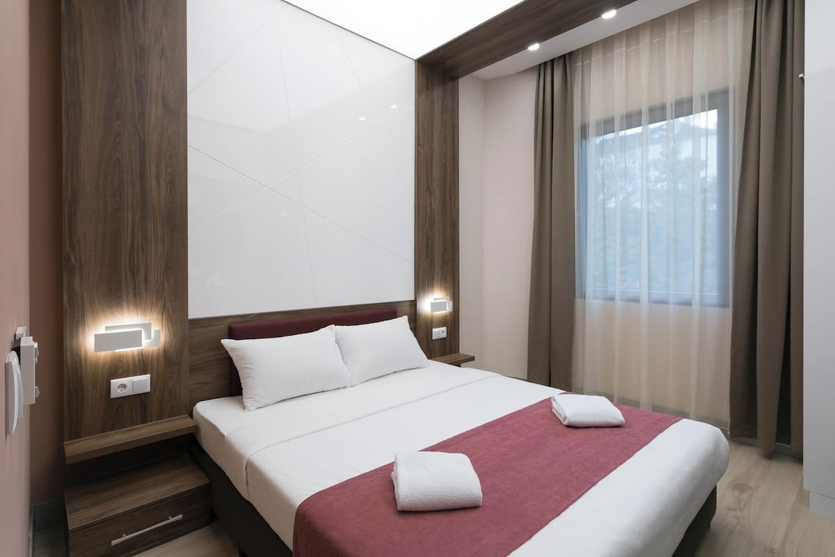 A modern hotel room with a large bed, wooden paneling, a view of trees through sheer curtains, and a nightstand with a lamp.