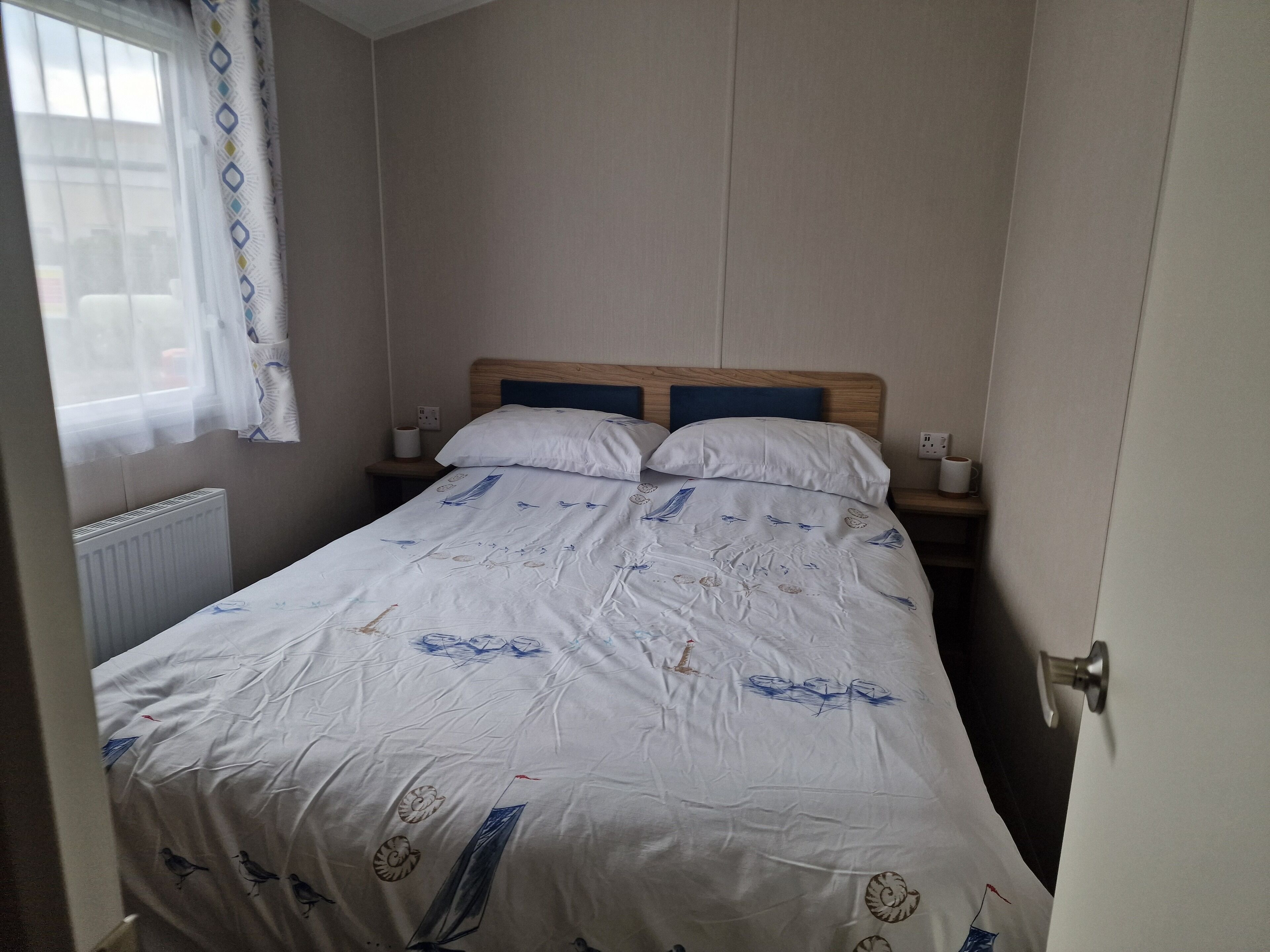 Ein kleines, ordentlich bezogenes Bett mit einem Holz-Kopfteil in einem kompakten Zimmer, das einen Heizkörper und ein mit Vorhängen versehenes Fenster hat.
