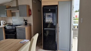 A modern kitchen with a stove, oven, and a black refrigerator. There is a dining table with chairs and a blue mat at the entrance. - Welcoming 3 Bedroom Caravan 2 Min Walk to Beach (Par)