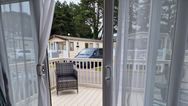 A view from a balcony with a wicker chair, a wooden deck, and a building with a car parked in front. - Welcoming 3 Bedroom Caravan 2 Min Walk to Beach (Par)