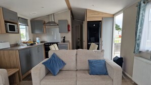 A compact living space with a kitchenette, a sofa with blue cushions, and a view of the outside. - Welcoming 3 Bedroom Caravan 2 Min Walk to Beach (Par)