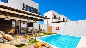 Un área de piscina moderna al aire libre con sillones de descanso, un comedor y un porche cubierto.