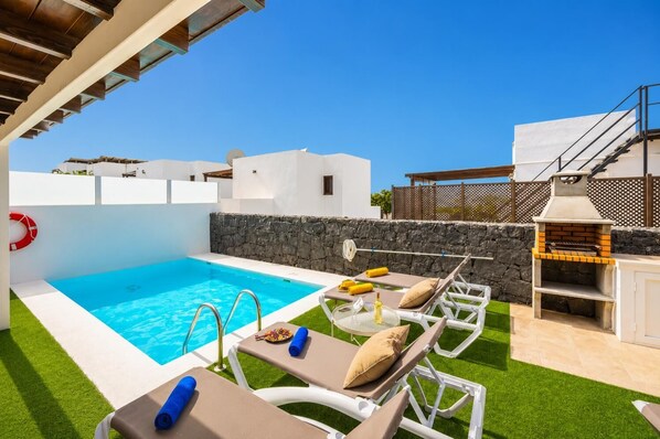 Un área de piscina exterior moderna con sillones de descanso, una parrilla y un muro de piedra.