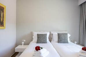 A hotel room with two beds, white linens, grey pillows, a bedside table with a lamp, and a painting on the wall.