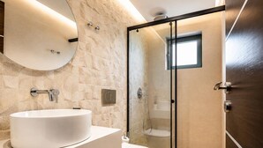 Ein modernes Badezimmer mit Steinwand, rundem Spiegel, weißem Waschbecken, Toilette und einer Duschkabine aus Glas.