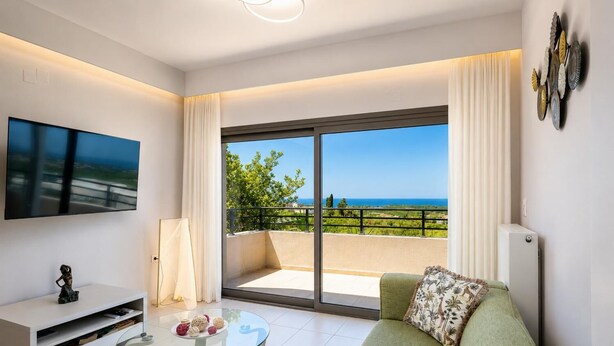 Ein modernes Wohnzimmer mit einem Glastisch, einer gepolsterten Couch mit einem floralen Kissen, einem Flachbildfernseher und einem Balkon mit Meerblick.