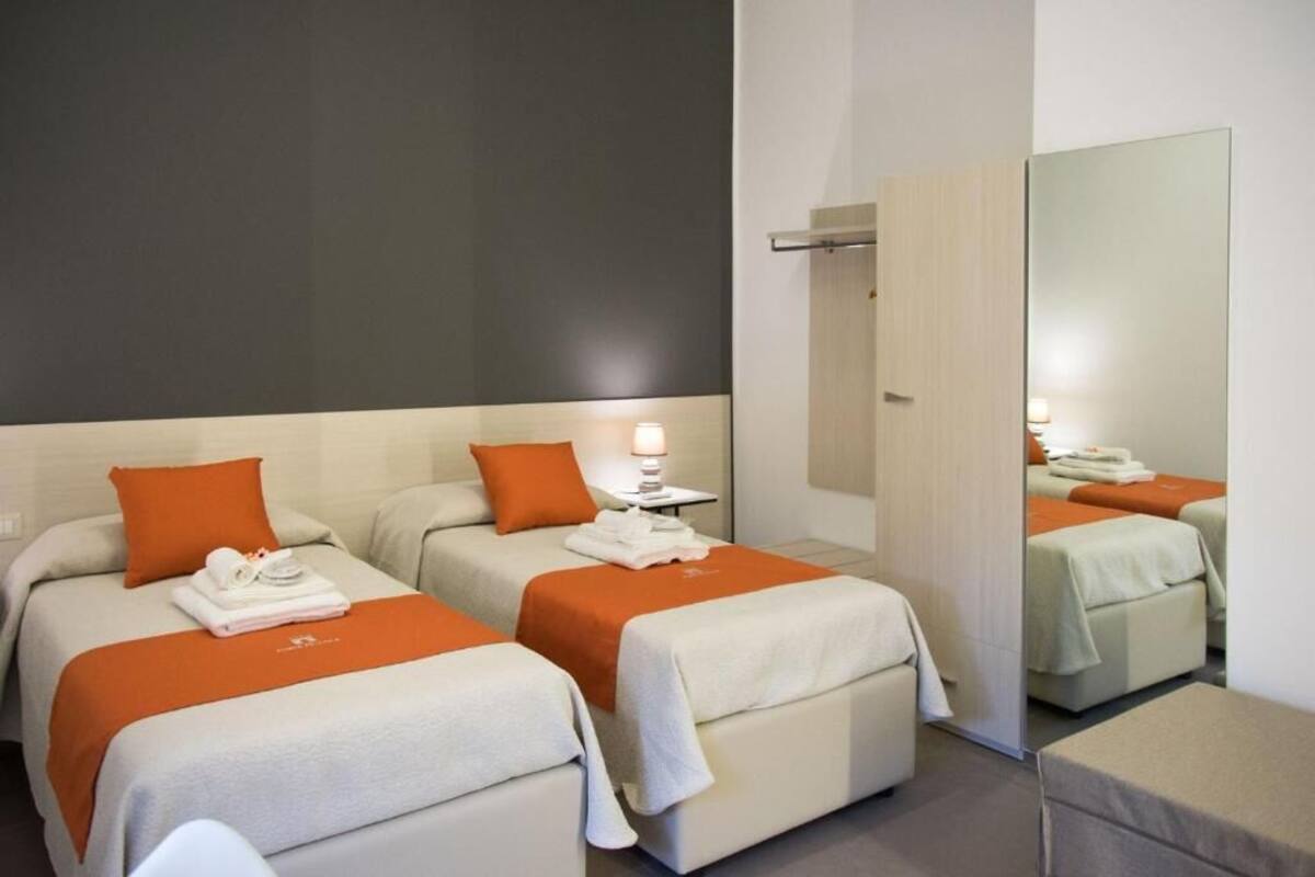 A hotel room with two beds, each with an orange blanket and pillows, a bedside table with a lamp, and a large mirror on the wall.