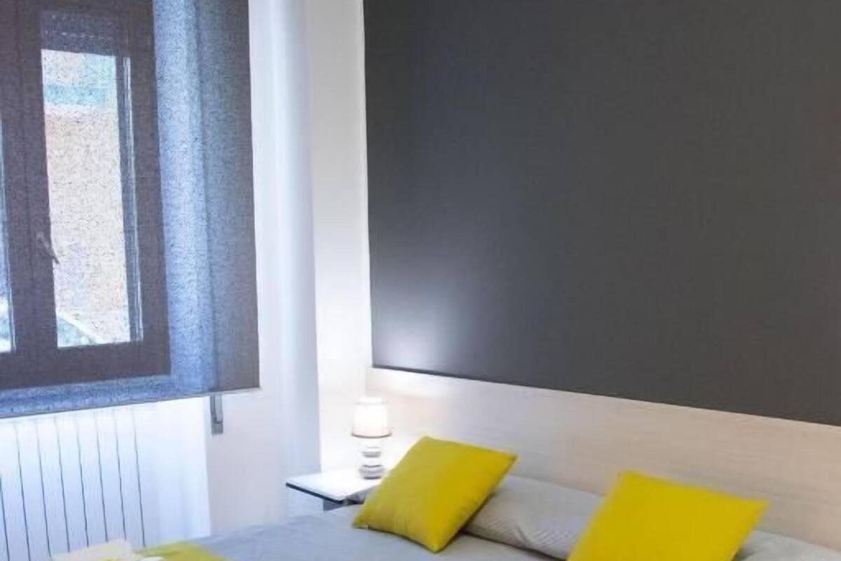 A modern hotel room with a large bed, a bedside table with a lamp, and a window with blinds.