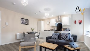 A modern living room with a grey sofa, a wooden coffee table, and a grey armchair. A kitchen is visible in the background with white cabinets and stainless steel appliances.