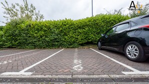 A parking area with a car parked, a hedge, and a paved surface.