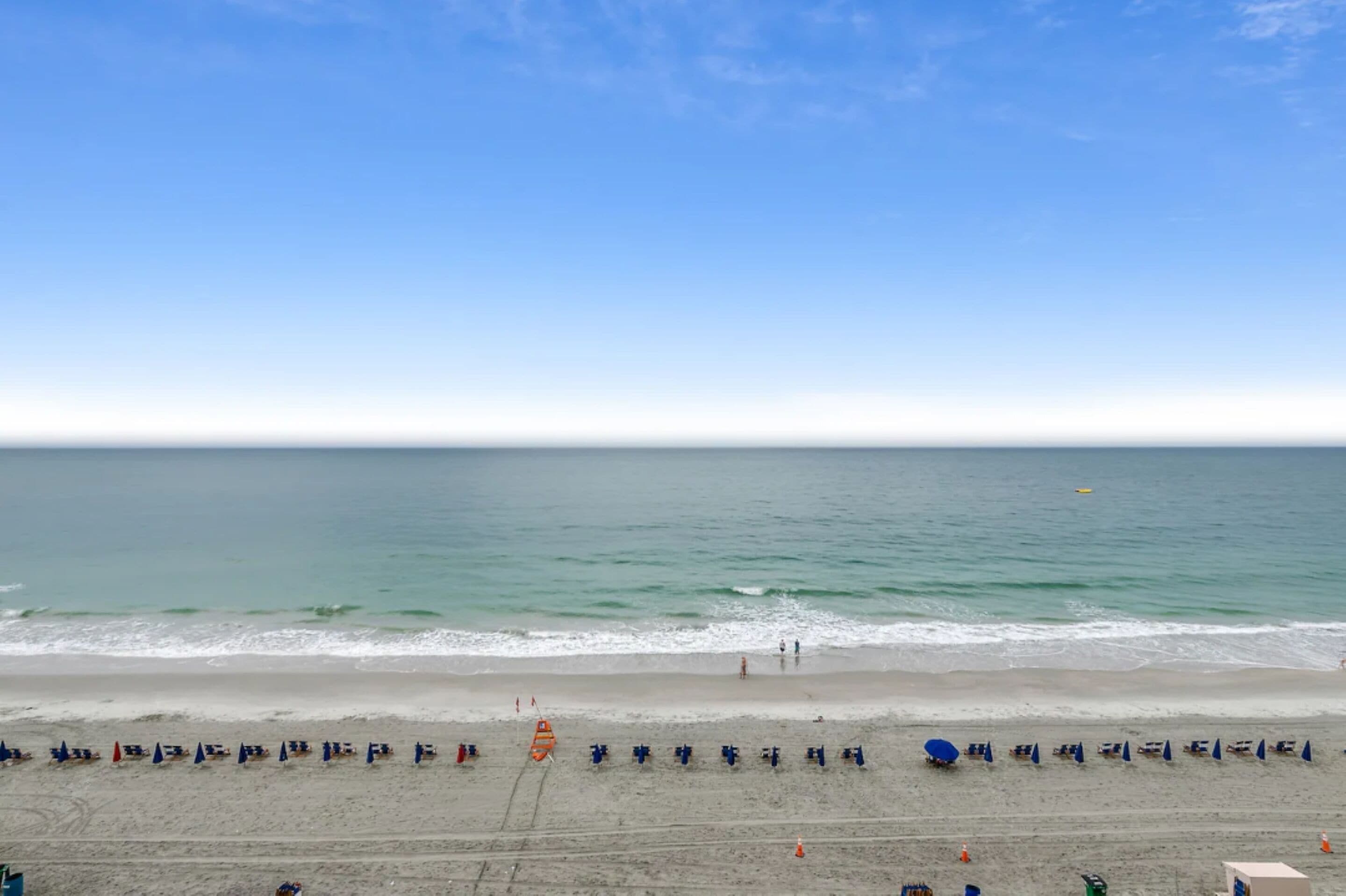 Une plage bordée de rangées de parasols et de chaises longues, un ciel dégagé et une mer calme.