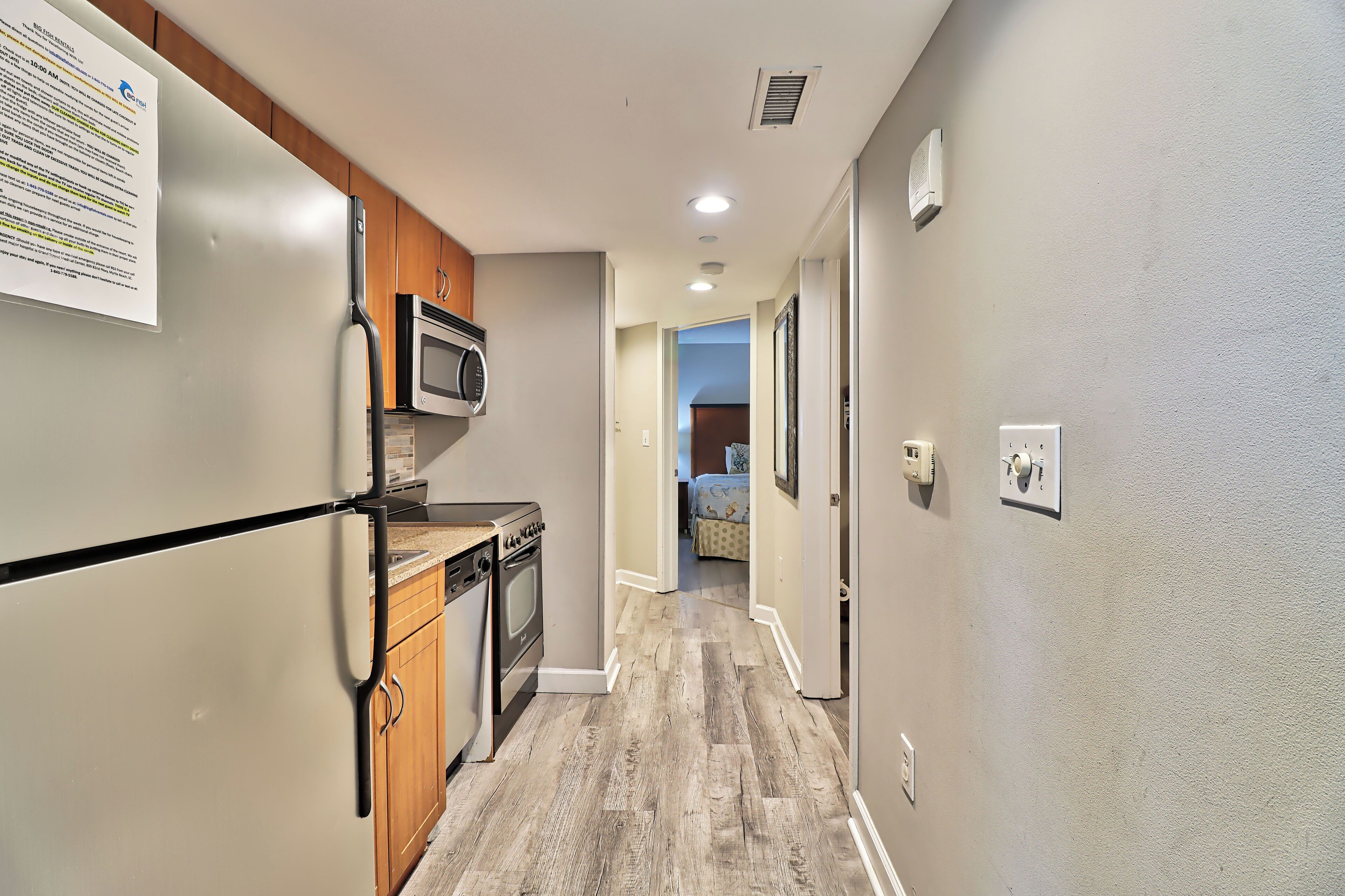 A narrow hallway with a kitchen area on the left, including a refrigerator, microwave, and stove. The hallway leads to a bedroom with a bed and a window.