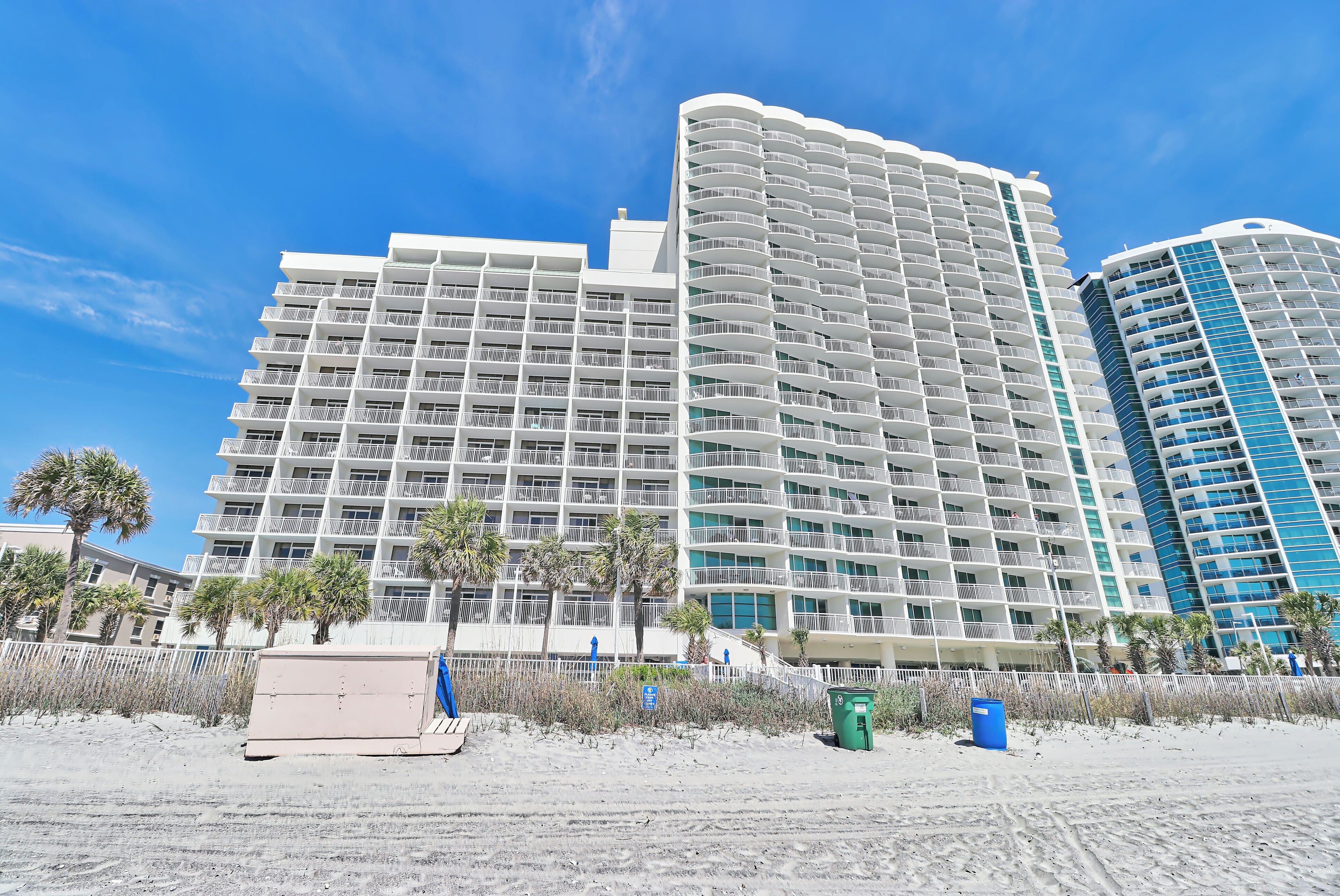 A high-rise hotel with multiple balconies, a sandy beach in the foreground, and palm trees.