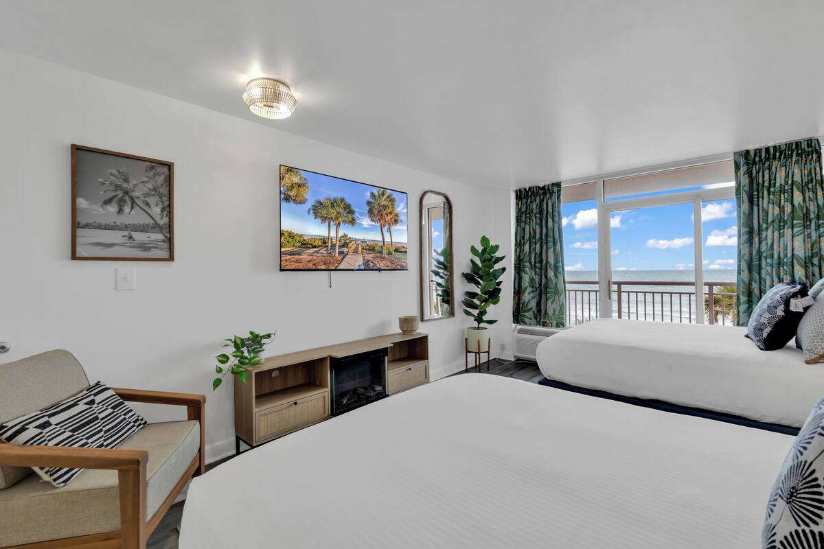 A modern hotel room with a large bed, a sofa, a fireplace, a view of the sea, and tropical-themed decor.
