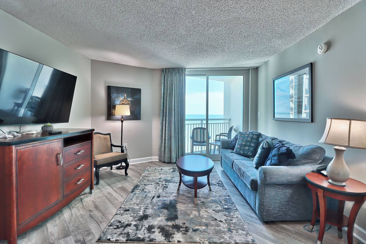 A modern hotel room with a large flat-screen TV, a sofa, a coffee table, a cabinet, and a view of the outside.