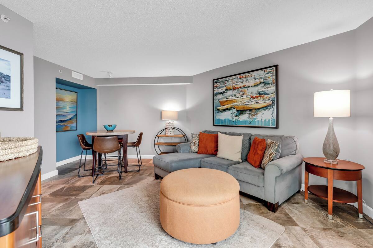 A modern living room with a grey sofa, round ottoman, and a dining area with a table and chairs.