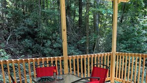 A wooden deck with a glass table and red chairs, surrounded by a wooden railing and dense forest. -  Tiny Cabin in the Laurels, with queen bed & queen sofa bed in wooded setting. (Marion)