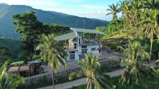 Um edifício de dois andares com uma varanda, cercado por palmeiras e montanhas ao fundo.