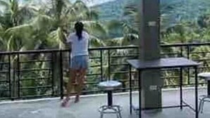 Uma pessoa em uma varanda, com vista para palmeiras e uma montanha.