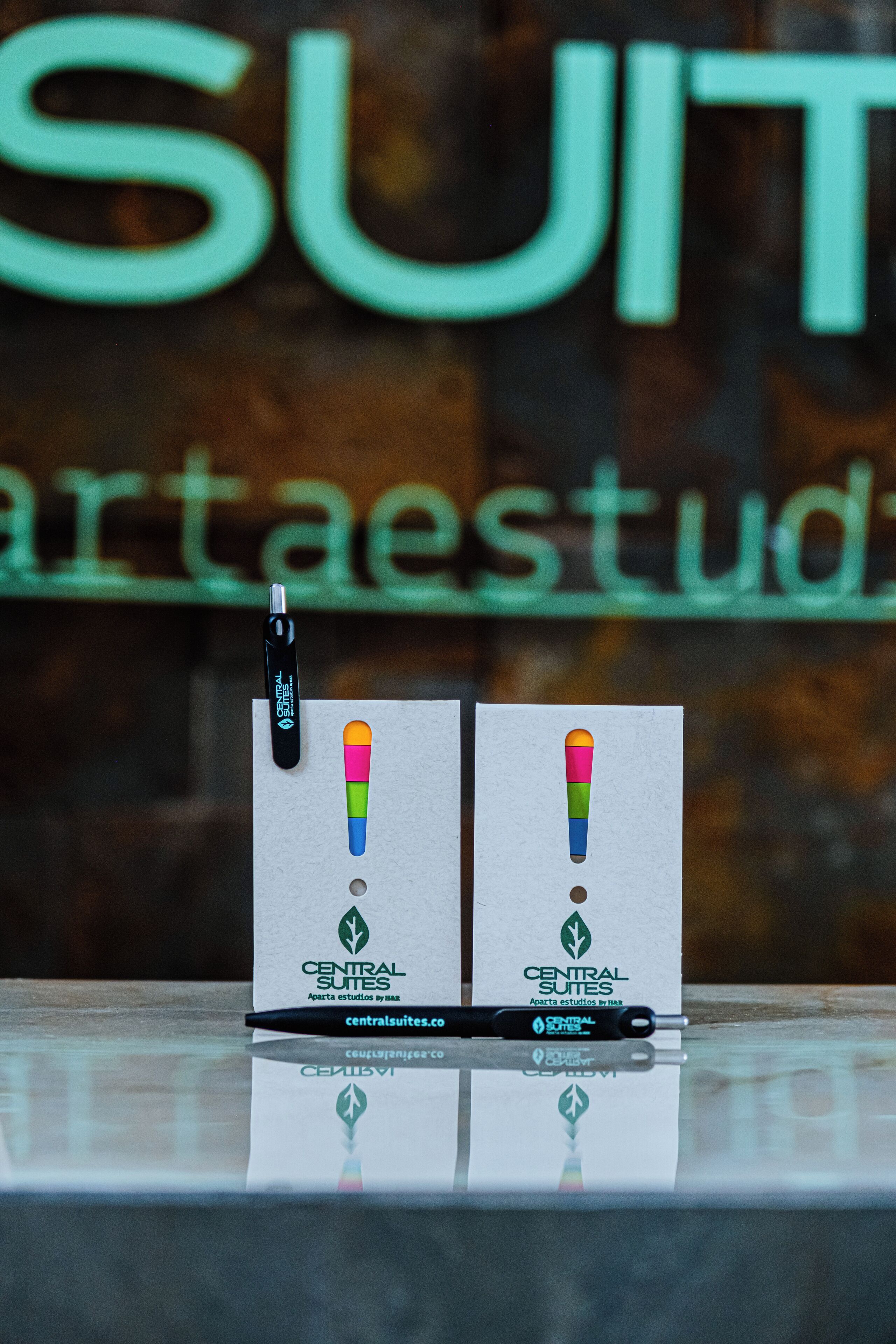 Una pluma y dos tarjetas con un diseño colorido sobre una superficie reflectante, con un cartel al fondo.