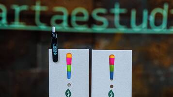 Una pluma y dos tarjetas con un diseño colorido sobre una superficie reflectante, con un cartel al fondo.