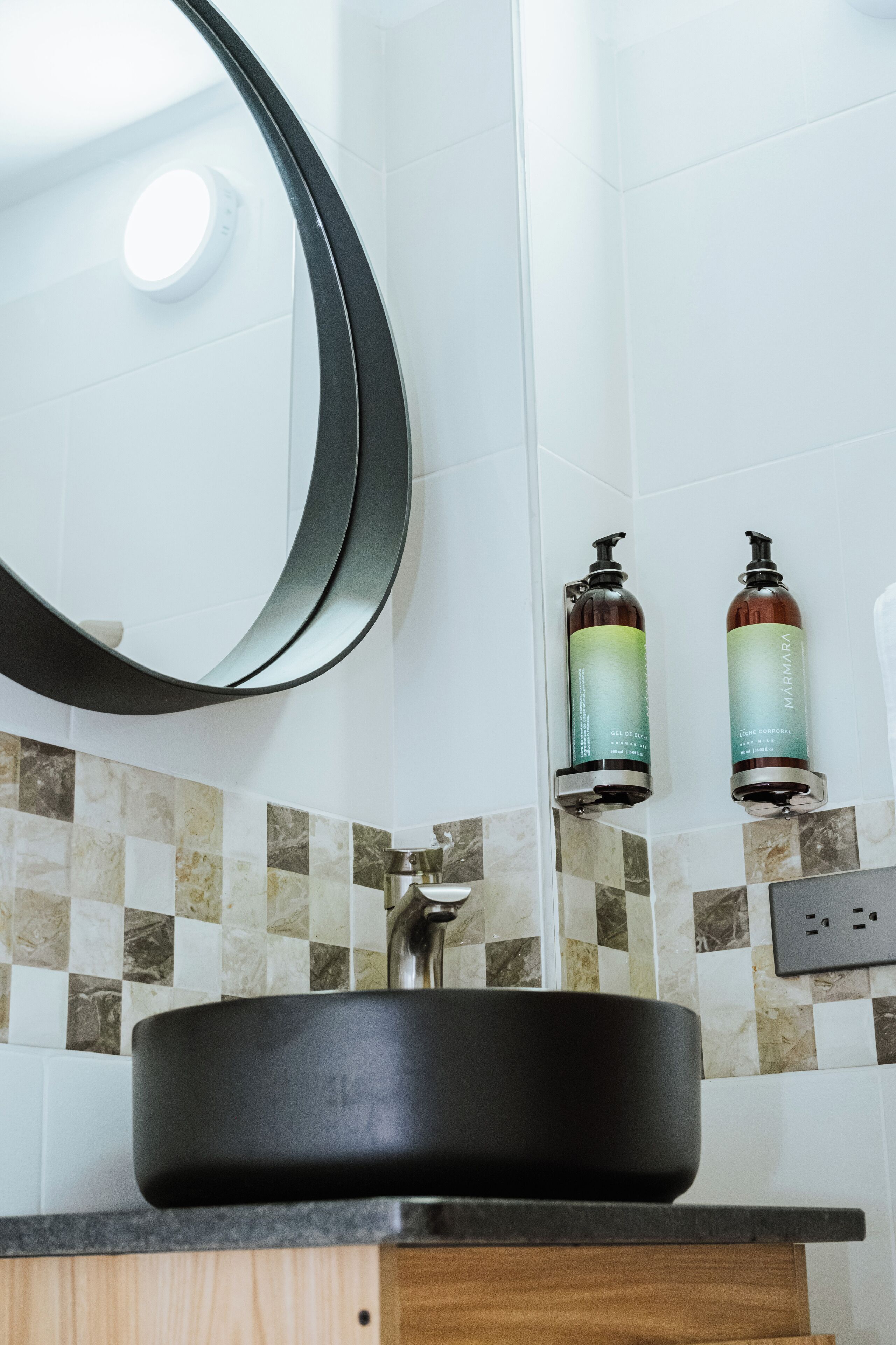 Une salle de bain avec un lavabo noir, un miroir rond et deux bouteilles de shampoing accrochées au mur.
