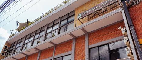 A multi-story building with a sign for "CENTRAL SUITES" indicating it is a hotel. The building has multiple balconies with plants, large windows, and a brick facade.