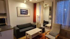 Ein modernes Wohnzimmer mit einer schwarzen Couch, einem kleinen Tisch, einem Sessel mit gestreifter Sitzkissen und einem gerahmten Bild an der Wand. Ein großes Fenster mit Vorhängen vervollständigt den Raum.