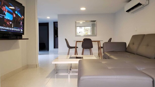 A modern living room with a flat-screen TV, a sofa, a dining table with chairs, and an air conditioning unit. - 2 bedroom apartment in the best area of Laureles. A/C. (Medellín)