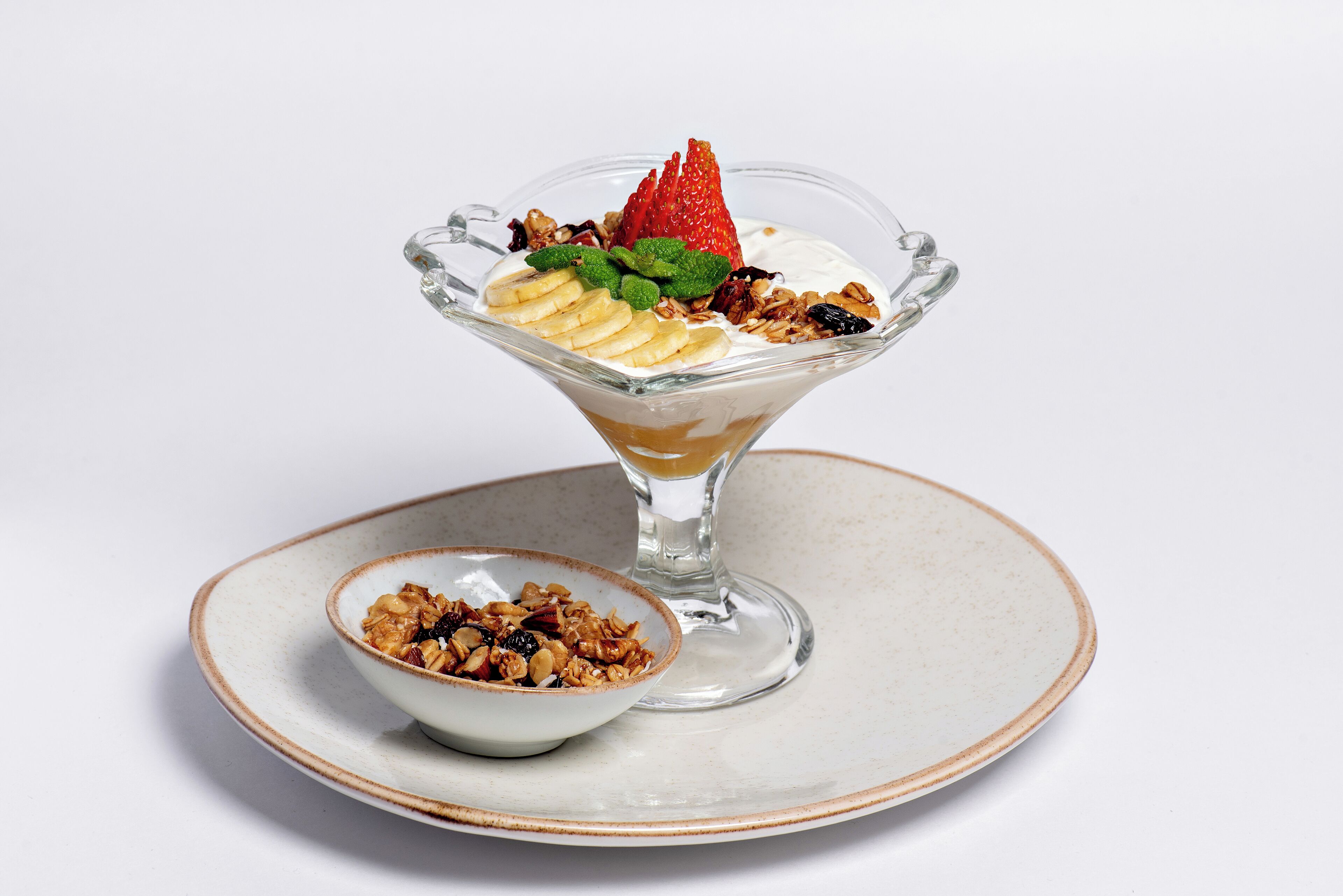 A dessert in a glass bowl on a plate, with a smaller bowl of granola beside it.