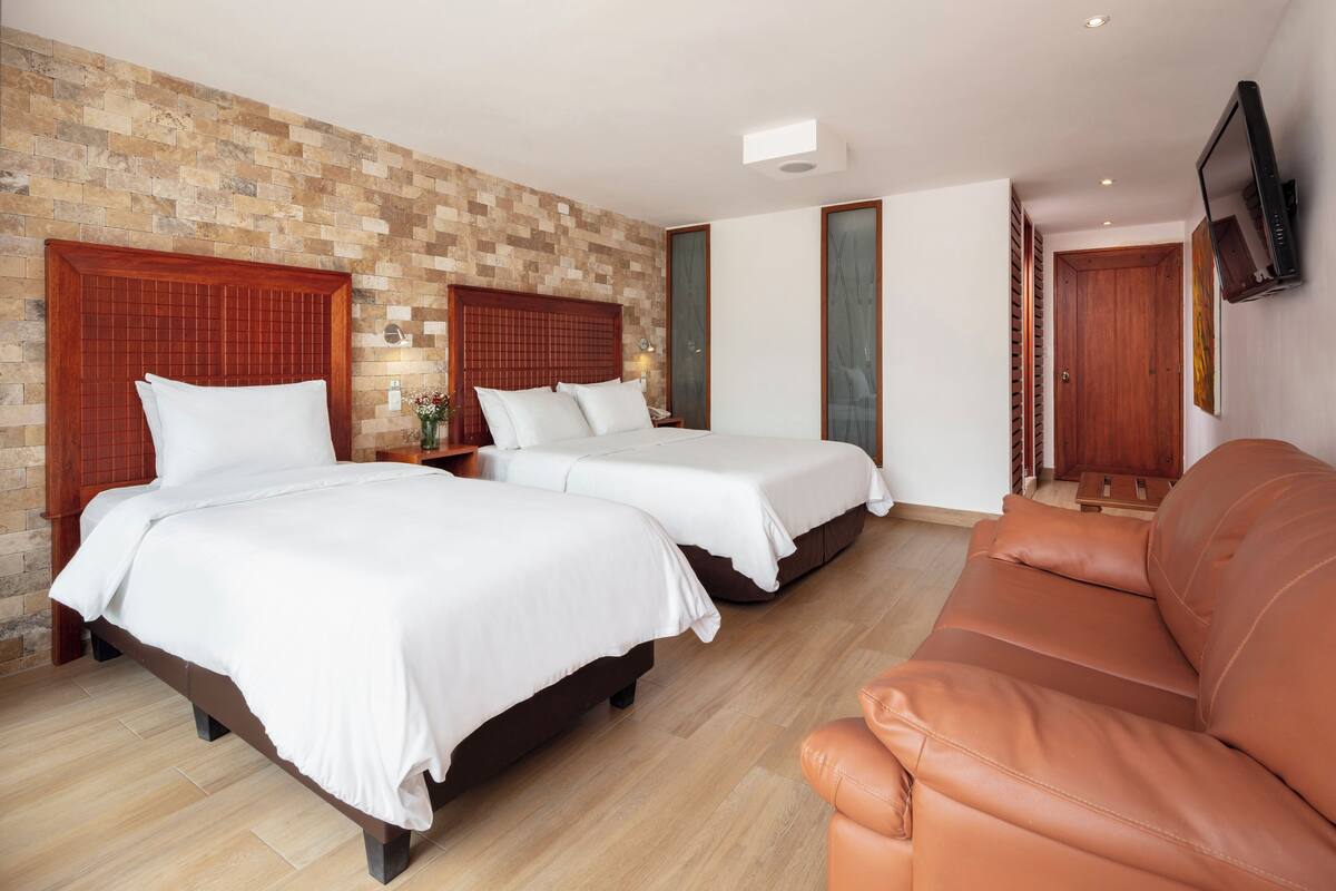 A hotel room with two beds, a brown leather sofa, and a brick-patterned wall.