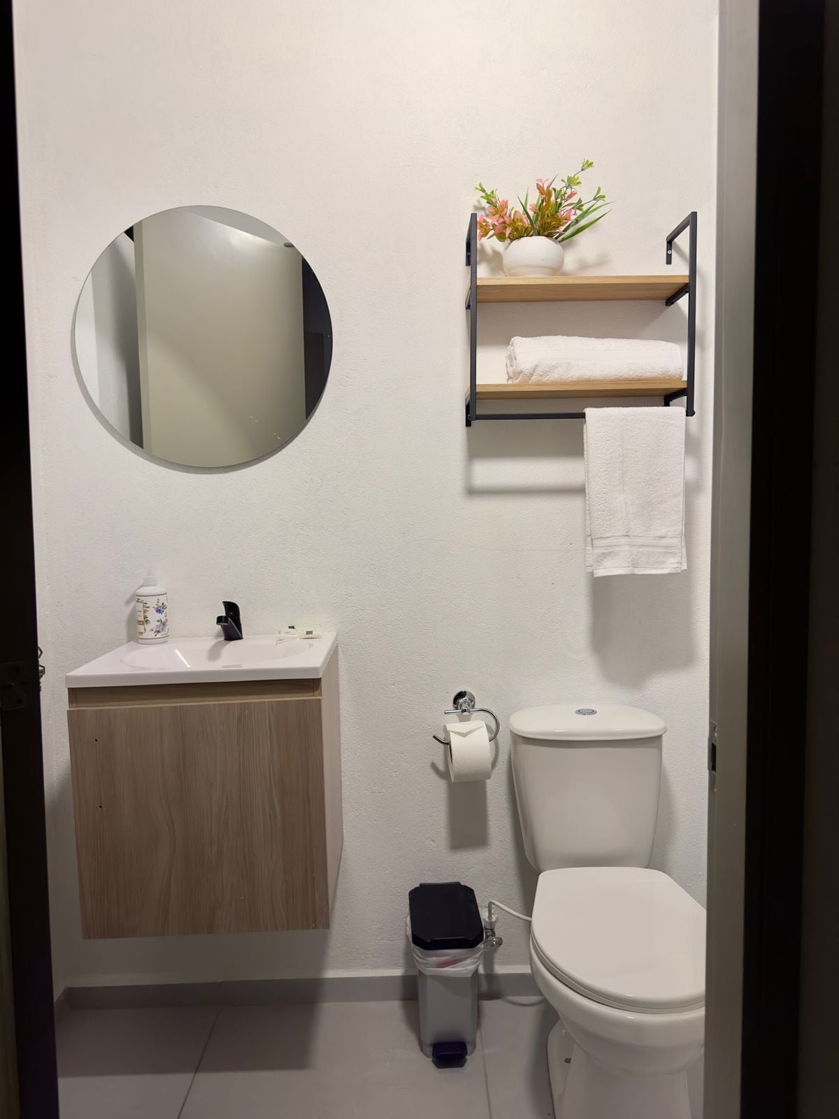 Un baño pequeño con un espejo redondo, un inodoro, un lavabo con mueble de madera y un cubo de basura.